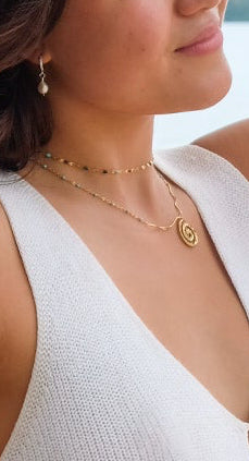 Woman in a white top and beige pants leaning against a tree on a beach wearing the Oceana Necklace.